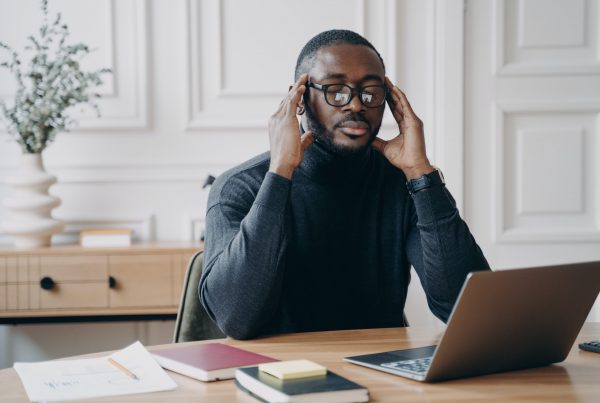 African male bookkeeper sits at desk in front of laptop eyes closed fingers gently touching temples